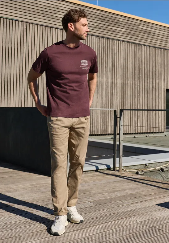 Een man staat op een houten terras, draagt een bordeaux T-shirt, beige broek en witte sneakers.