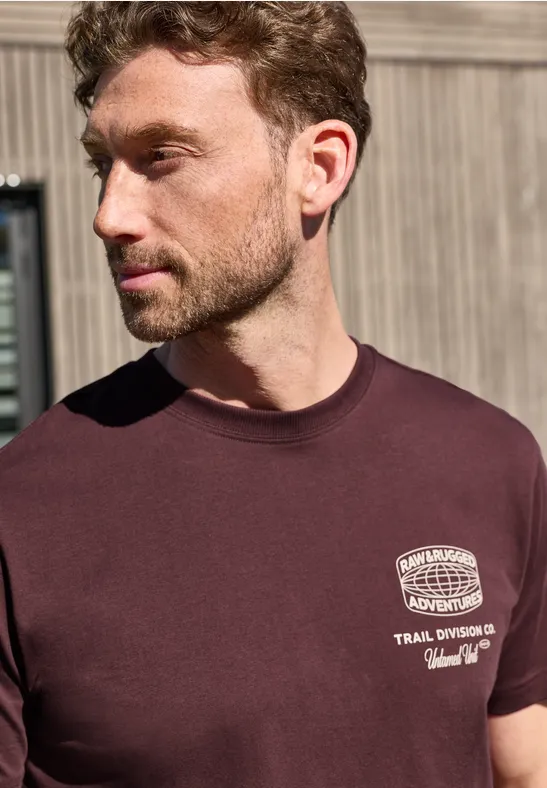Een jonge man draagt een bordeauxrood t-shirt met witte opdruk en staat voor een houten wand.