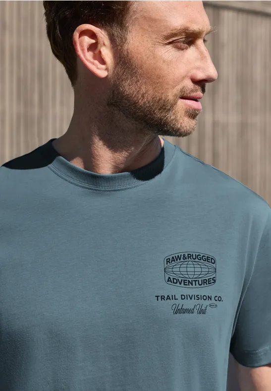 Ein seitliches Porträt eines Mannes, der ein türkisfarbenes T-Shirt mit Abenteuer-Print trägt.