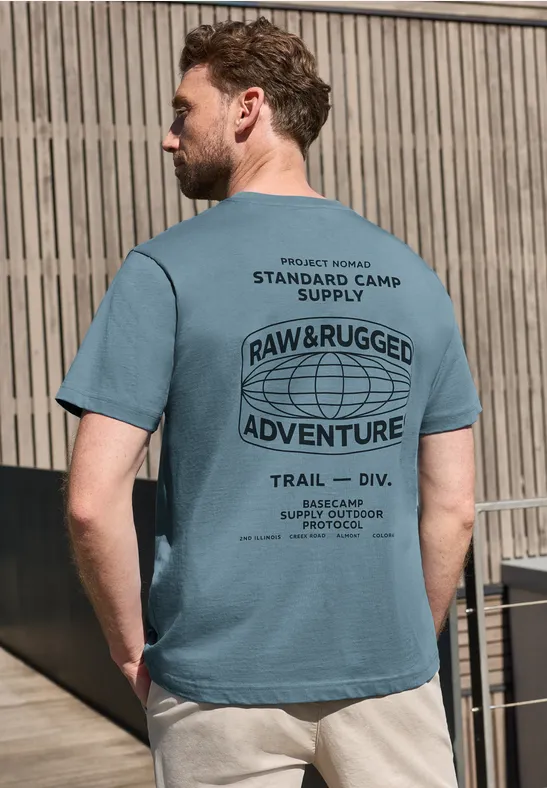 Mann mit türkisfarbenem T-Shirt und beigen Shorts lehnt an Holzlattenzaun, Rückansicht.