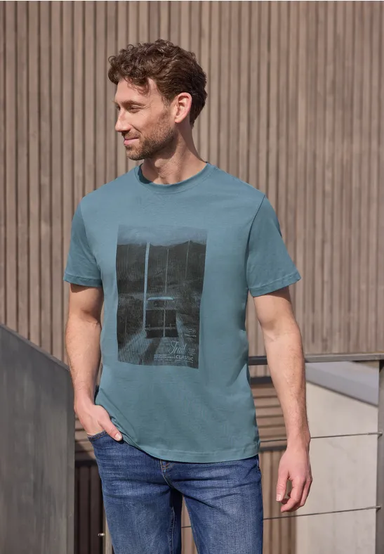 Ein Mann in türkisfarbenem T-Shirt mit Print und Jeans steht lässig vor einer holzverkleideten Wand.