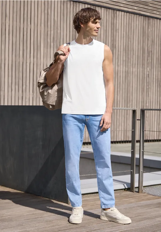 Een man staat voor een houten gevel, draagt een wit mouwloos shirt, lichte spijkerbroek, beige sneakers en een tas.