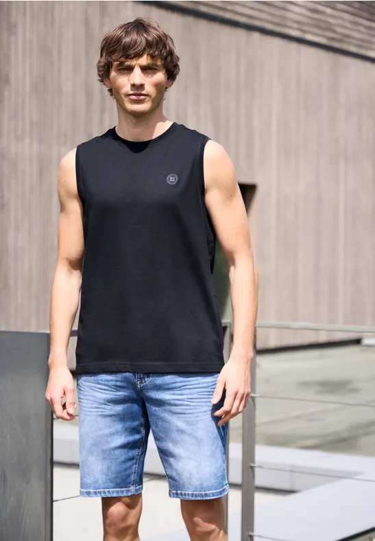 Junger Mann in schwarzem ärmellosem Shirt und blauen Jeans-Shorts vor moderner Fassade.