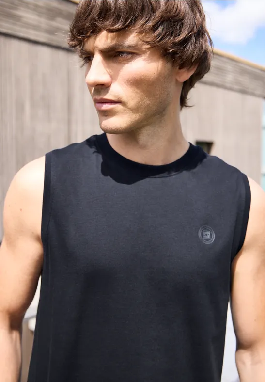 Ein athletischer Mann in einem schwarzen, ärmellosen Shirt steht im Freien, Gesicht unscharf.