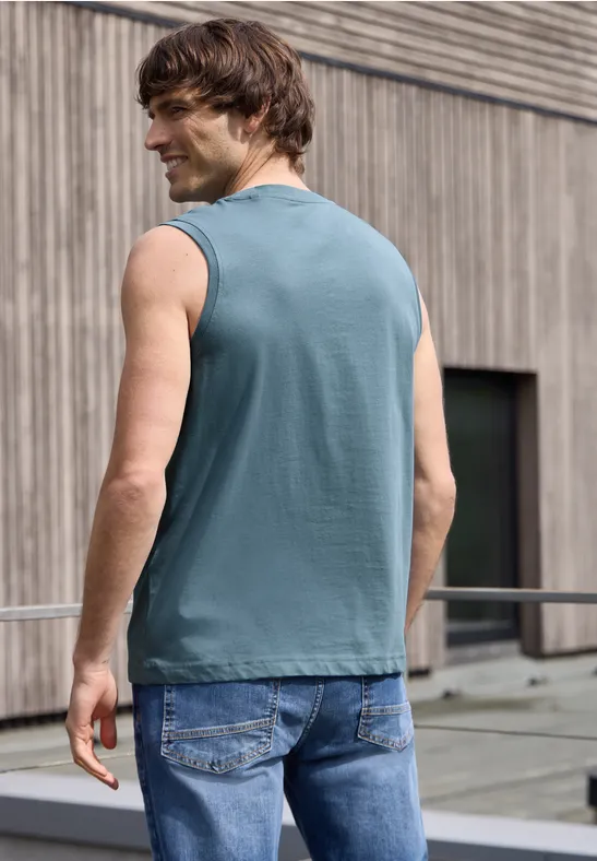 Rückenansicht eines Mannes in ärmellosem Shirt und Jeans auf einem Dachbalkon vor einer Holzfassade.