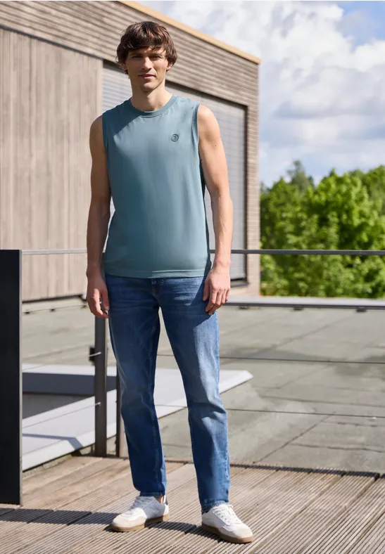 Ein Mann mit ärmellosem Shirt und Jeans steht auf einer Holzterrasse vor modernem Gebäude und Grün.
