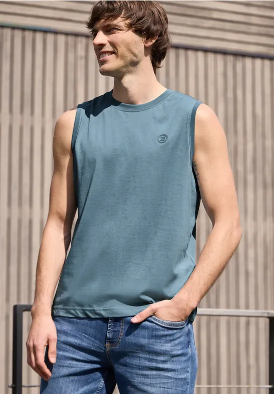 Ein junger Mann trägt ein ärmelloses Shirt und Jeans, steht entspannt vor einer Holzfassade.