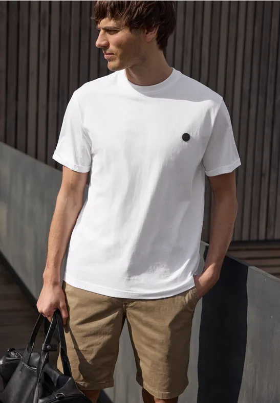 Mann in weißem T-Shirt und beigen Shorts steht mit Tasche in Hand vor dunkler Holzfassade.
