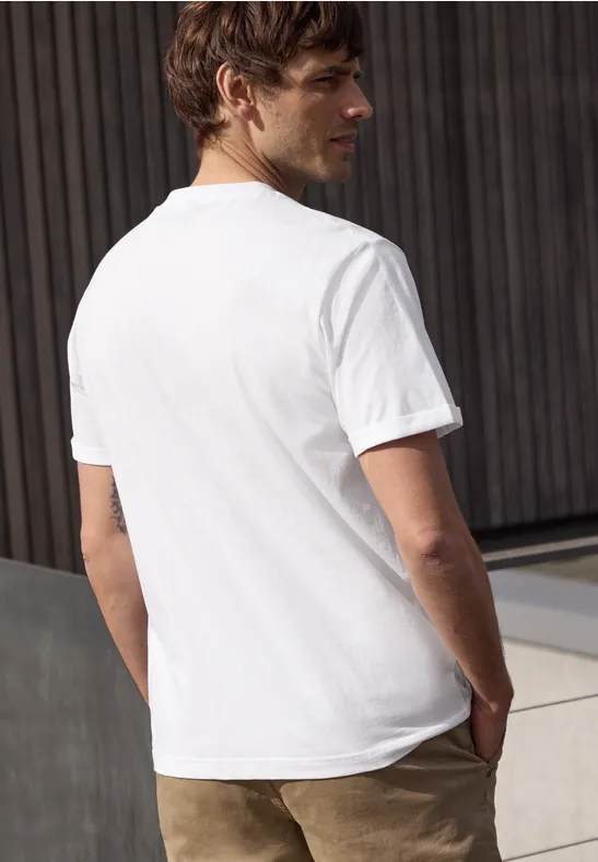 Mann von hinten in weißem T-Shirt und brauner Hose vor Holzwand.