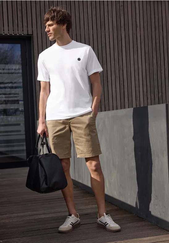 Ein Mann in weißem T-Shirt, beigen Shorts und Sneakers hält eine schwarze Tasche vor einer modernen Fassade.