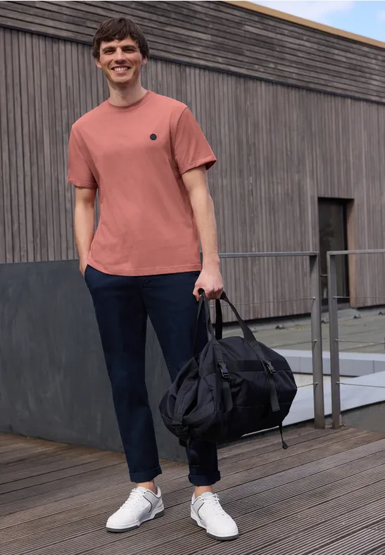 Ein Mann in rotem T-Shirt und dunkler Hose trägt eine schwarze Reisetasche auf einer Holzterrasse.