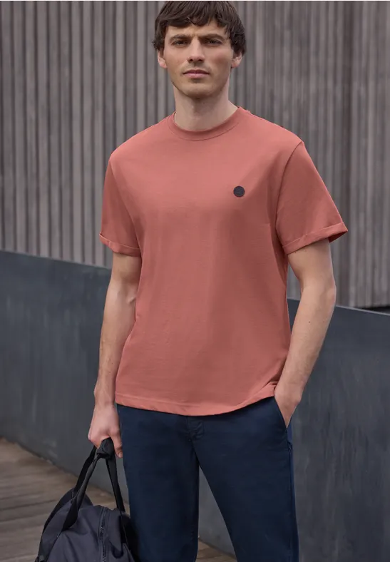 Ein Mann in rostfarbenem T-Shirt und dunkler Hose hält vor Holzvertäfelung eine schwarze Tasche.