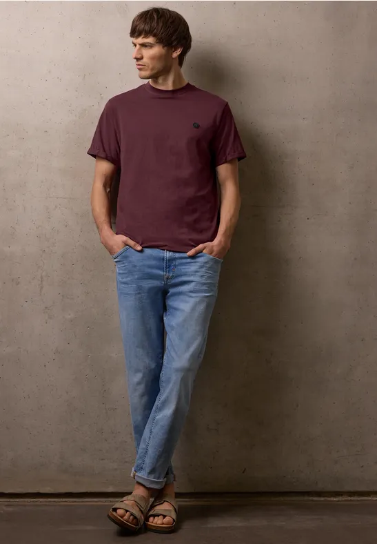 Mann in bordeauxfarbenem T-Shirt, blauen Jeans und Sandalen lehnt lässig an Wand.