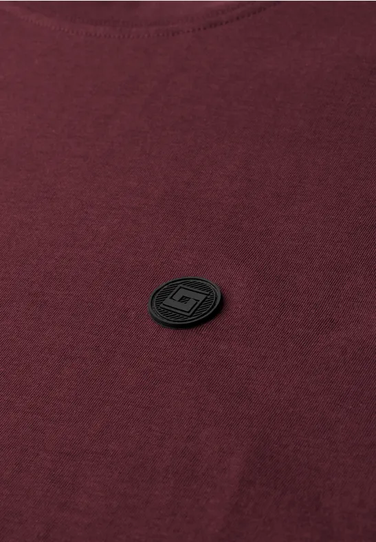Ein dunkelrotes T-Shirt mit einem schwarzen, runden Emblem auf der Brust.