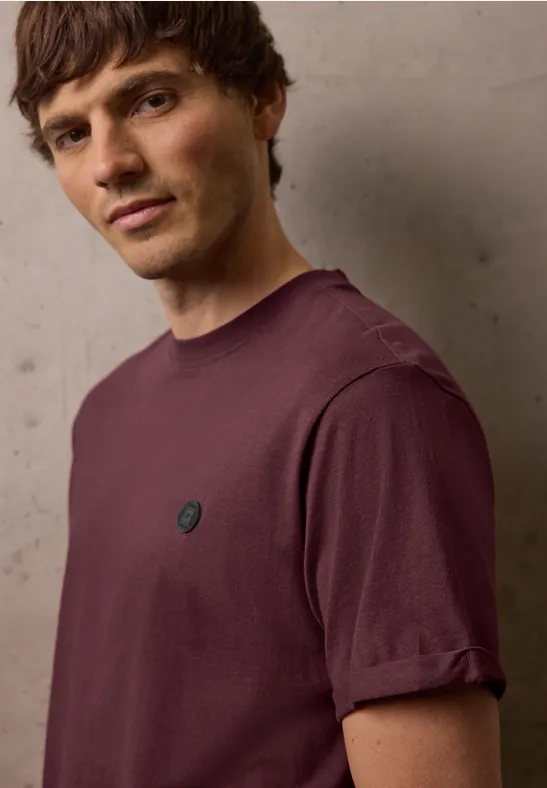 Ein Mann mit hochgekrempeltem Ärmel trägt ein schlichtes violettes T-Shirt mit kleinem Logo auf der Brust.