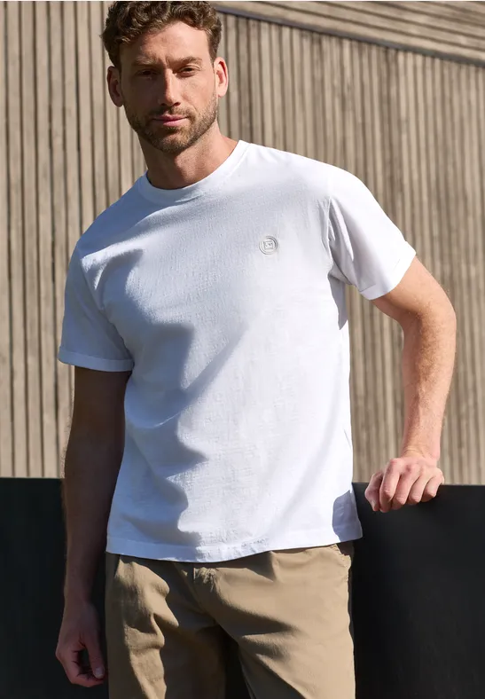 Een man in een wit T-shirt en beige broek staat voor een houten gevel.