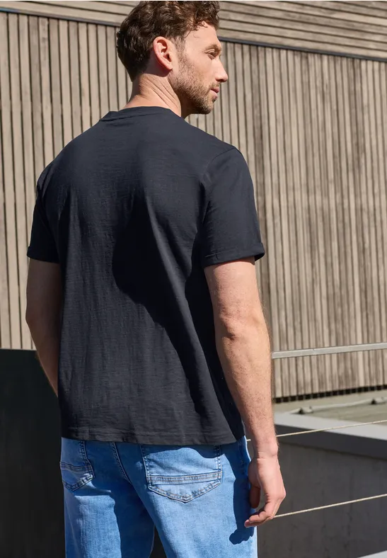 Ein Mann steht mit schwarzem T-Shirt und blauen Jeans vor einer Holzverkleidung.