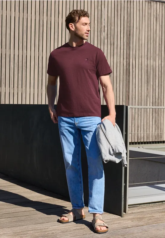 Ein Mann in burgunderfarbenem T-Shirt und blauen Jeans hält eine graue Jacke und Laptophülle.
