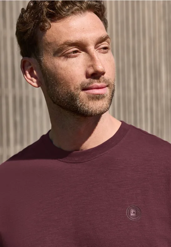 Ein Mann mit verschwommenem Gesicht trägt ein dunkelrotes T-Shirt vor einer Holzlattenwand.