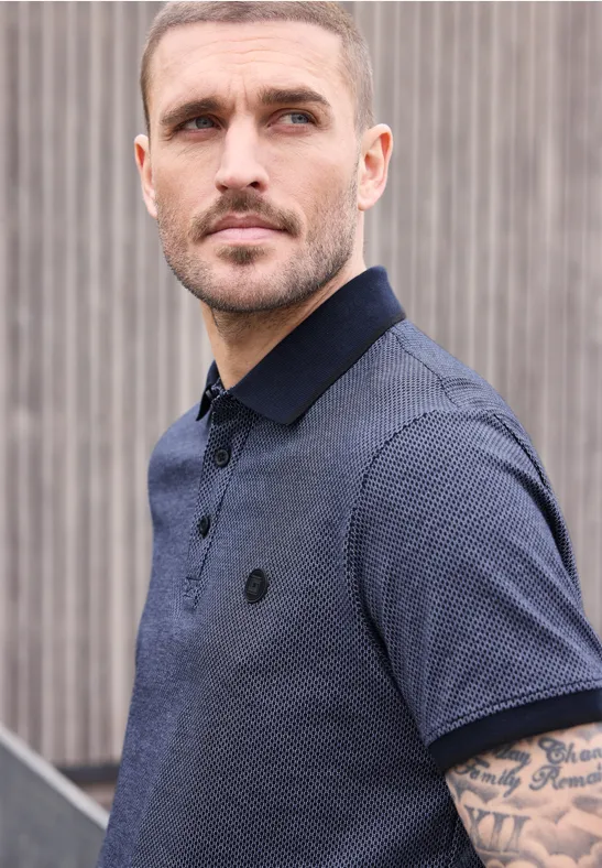 Ein Mann in blauem Poloshirt steht vor einer Holzwand, sichtbarer Unterarm mit Tattoo.