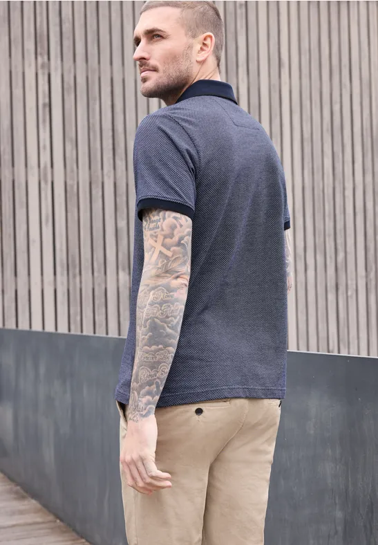 Ein tätowierter Mann trägt ein blaues Polohemd und beige Hose und steht vor einem Holzlattenzaun.