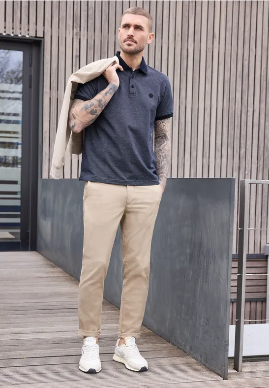 Ein tätowierter Mann in Poloshirt, beigen Chinos und weißen Schuhen trägt eine Jacke über der Schulter.