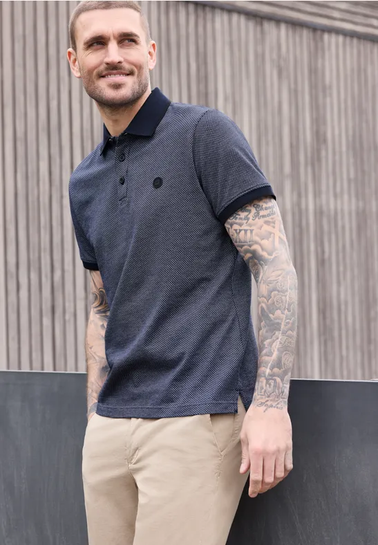 Ein tätowierter Mann trägt ein dunkelblaues Poloshirt und helle Hose vor einer Holzfassade.