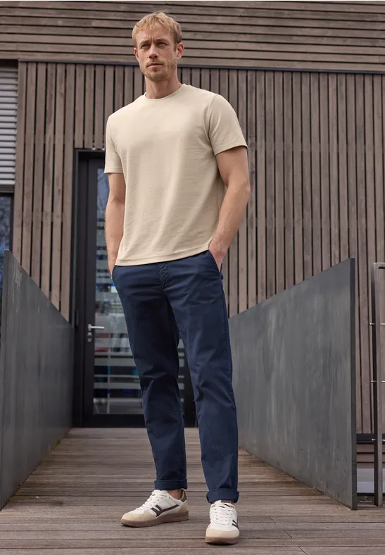 Ein Mann steht vor einer modernen Holzwand und trägt ein beiges T-Shirt, blaue Hose und weiße Sneaker.