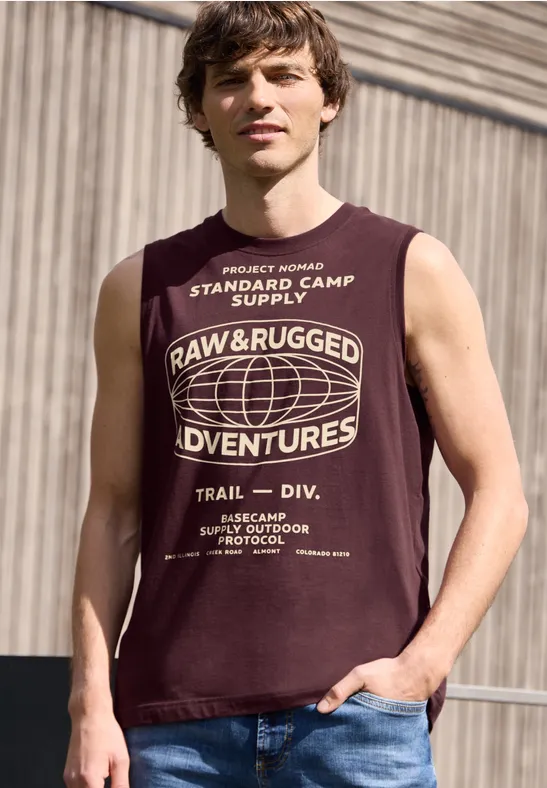 Ein Mann in ärmellosem, dunkelrotem T-Shirt mit weißem Aufdruck 'Raw & Rugged Adventures' vor Industriehintergrund.