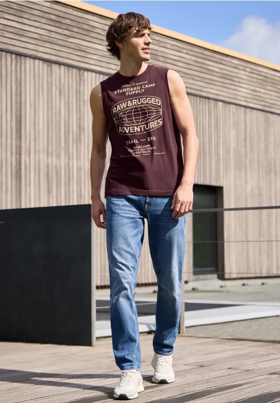 Ein junger Mann in ärmellosem Shirt und Jeans läuft auf einer Holzterrasse vor modernem Haus.