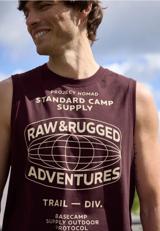 Ein Oberkörper in ärmellosem, dunkelrotem T-Shirt mit dem Aufdruck "RAW & RUGGED ADVENTURES" und Globus.
