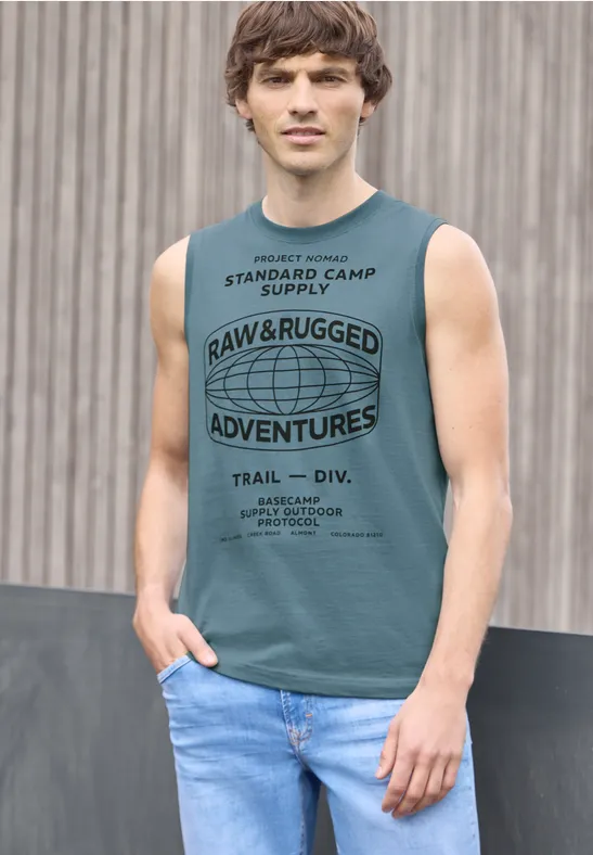 Ein junger Mann in ärmellosem grünen Shirt mit Aufdruck und blauen Jeans vor einer Holzpaneelwand.
