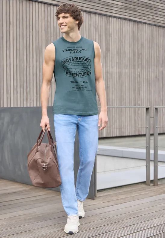 Ein Mann in Jeans und ärmellosem Shirt trägt eine Tasche und läuft auf einem Holzsteg vor einer Betonwand.