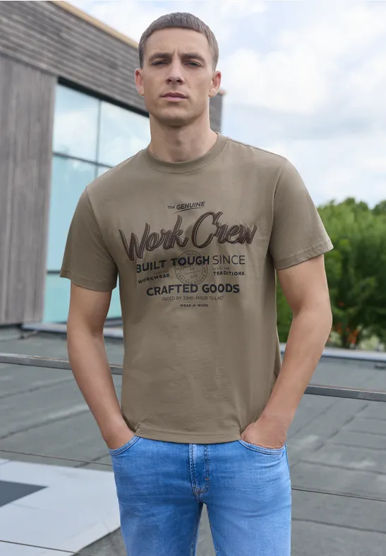 Ein junger Mann steht auf einer Dachterrasse, trägt ein khakifarbenes T-Shirt mit Aufdruck und eine blaue Jeans.