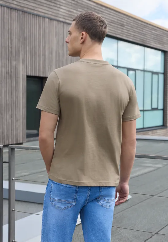 Ein Mann in Beige-T-Shirt und Jeans steht auf einer Dachterrasse, schaut auf moderne Glasfassade.