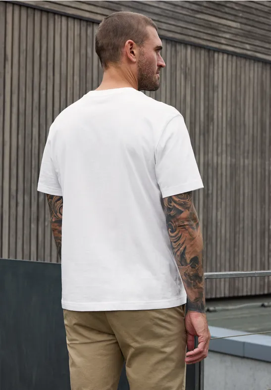 Tätowierter Mann in weißem T-Shirt und beiger Hose steht seitlich vor einer Holzwand.