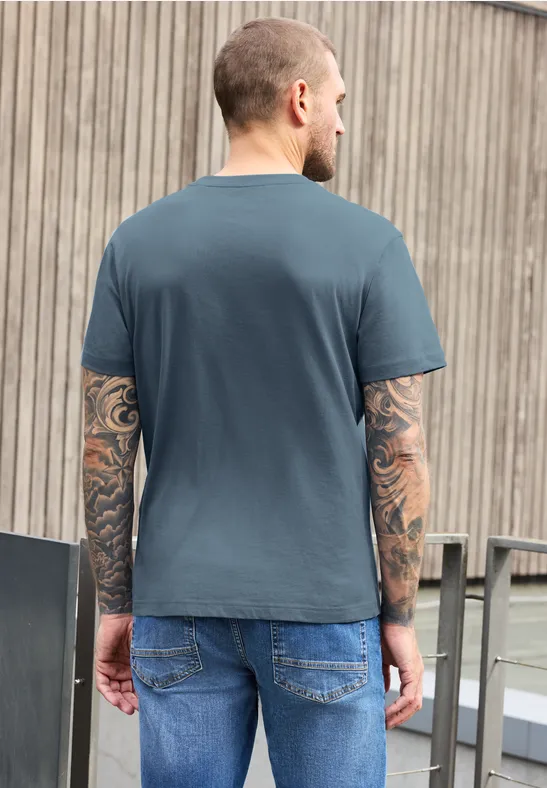 Ein Mann mit Tattoos auf beiden Armen, trägt graues T-Shirt und Jeans, steht Rücken zur Kamera vor einer Holzfassade.