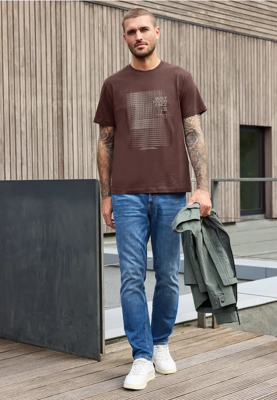Ein Mann mit braunem T-Shirt, Jeans und weißen Sneakern steht auf einer Holzterrasse und hält eine grüne Jacke.