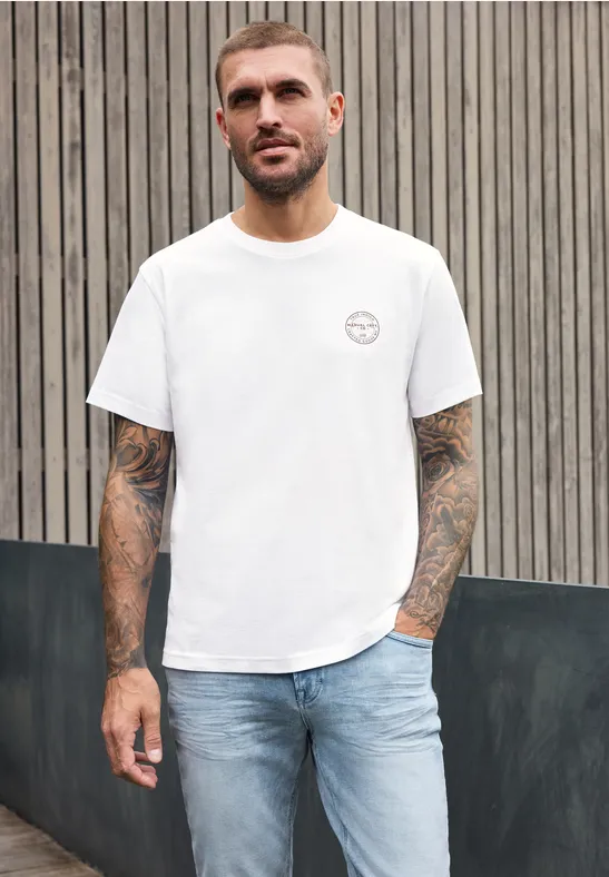 Ein tätowierter Mann trägt ein weißes T-Shirt und Jeans und steht vor einer hölzernen Wand.