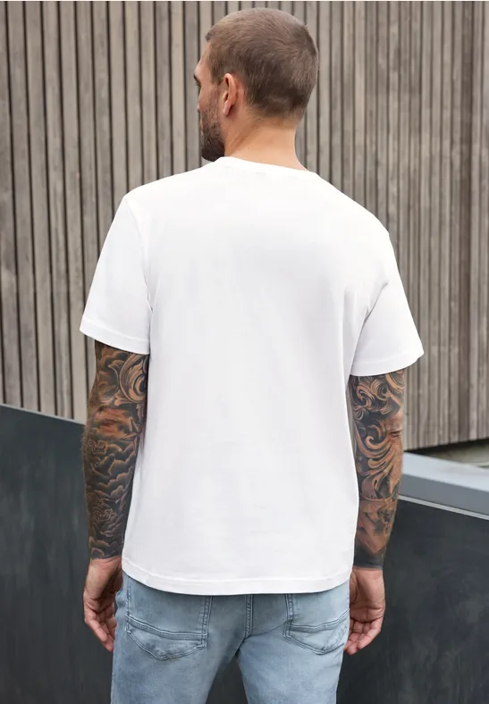 Ein tätowierter Mann in weißem T-Shirt und Jeans, Rückenansicht vor Holzwand.
