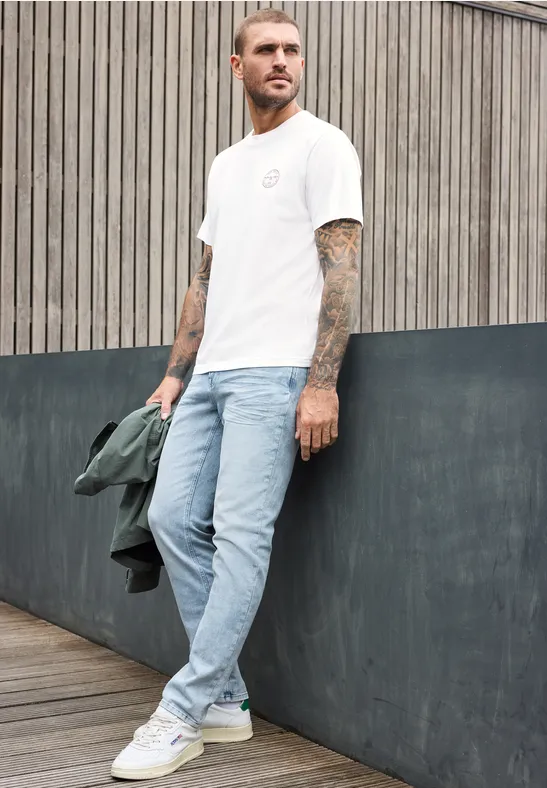Ein tätowierter Mann lehnt lässig an einer Wand, trägt Jeans, weißes T-Shirt und Sneakers und hält eine Jacke.