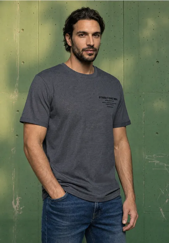 Ein Mann mit dunklem Haar und Bart trägt ein graues T-Shirt und Jeans vor einem grünen Hintergrund.