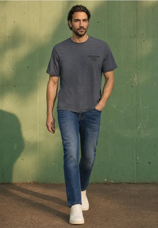 Ein Mann in grauem T-Shirt, Jeans und weißen Sneakers geht vor einer grünen Wand spazieren.