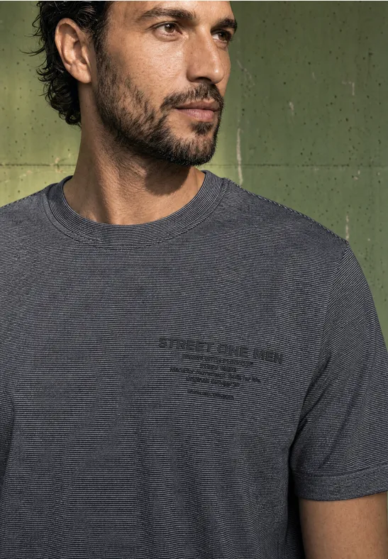 Mann mit braunem Haar und Bart trägt graues T-Shirt, schaut seitlich vor grünem Hintergrund.