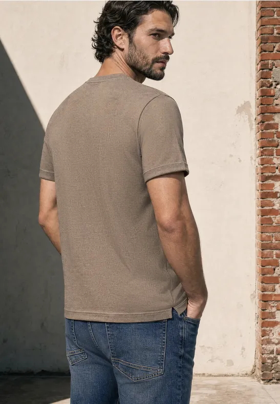 Een man staat met de rug naar de camera, draagt een beige t-shirt en spijkerbroek en kijkt over zijn schouder.