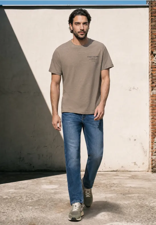 Een man in een T-shirt, spijkerbroek en sneakers loopt langs een zonnige muur.