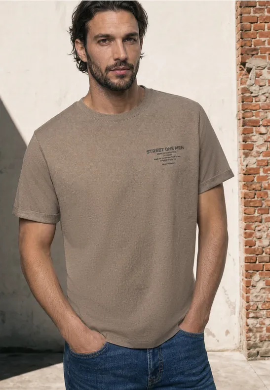 Man in beige T-shirt en blauwe jeans met handen in de zakken, staand tegen een bakstenen muur.