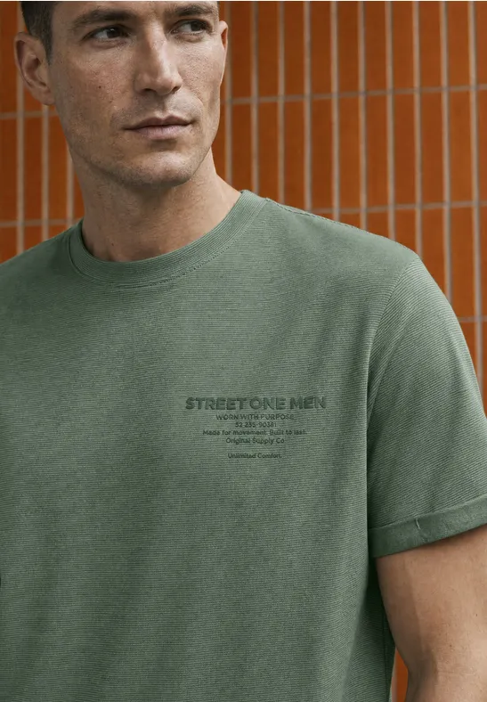 Man in groen T-shirt met klein logo, staat voor oranje betegelde muur.