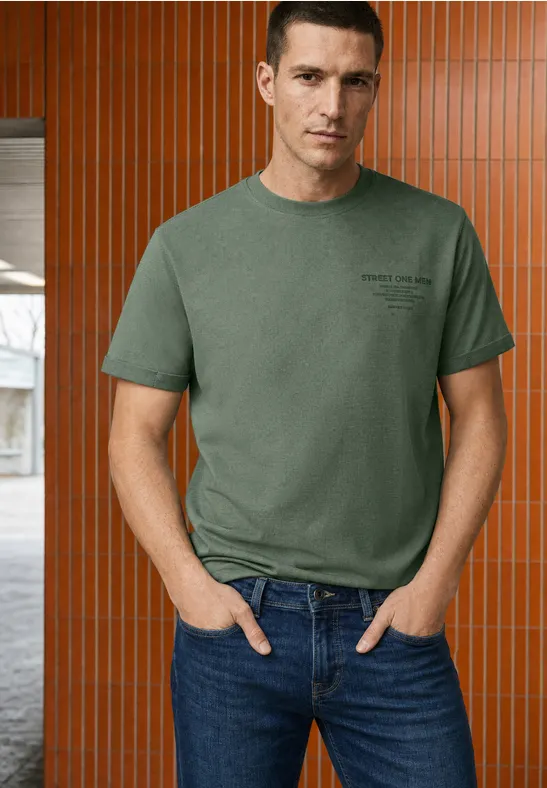 Een man in een groen T-shirt en jeans staat voor een bruin-oranje wand met verticale panelen.