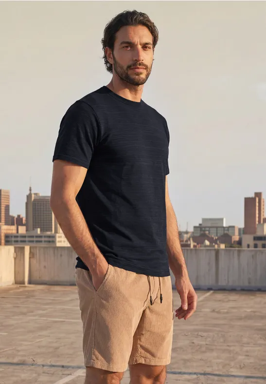 Ein Mann in schwarzem T-Shirt und beigen Shorts steht auf einem Dach und blickt in die Kamera vor Stadtkulisse.